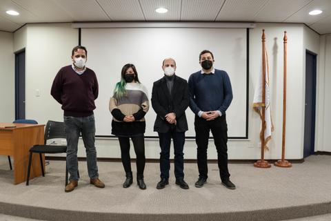 Cuatro personas posan en un evento en una sala con una presentación detrás de ellos.