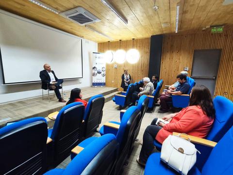 Una sala de reuniones con personas sentadas y un orador al frente.