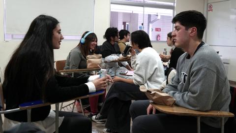 Un grupo de jóvenes conversando en un aula mientras algunos están sentados en escritorios.