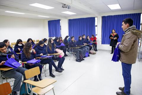 Un grupo de estudiantes está sentado en un aula mientras un presentador les habla y les entrega bolsas.