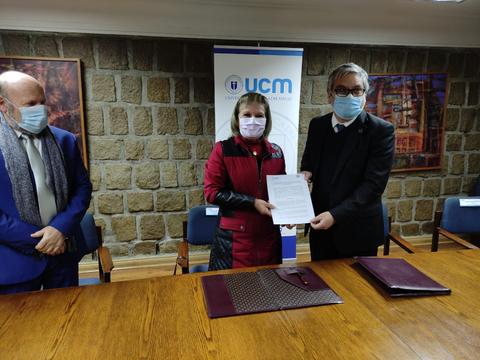 En la imagen se observa a dos personas firmando un documento en un evento formal, mientras que un tercer individuo las observa.