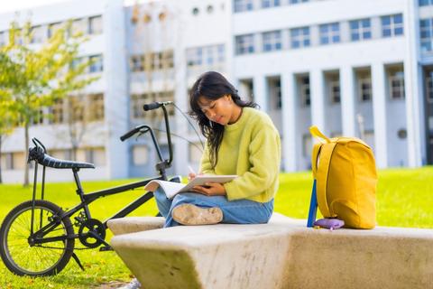 Una persona está estudiando en un parque, sentada sobre una piedra con su bicicleta al lado.
