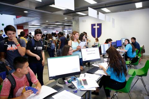 La imagen muestra un grupo de estudiantes en un ambiente académico, algunos en la fila y otros utilizando computadoras en un espacio de trabajo compartido.
