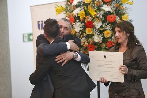 Un grupo de hombres se abrazan en un evento de entrega de premios mientras una mujer sostiene un certificado y hay flores de fondo.