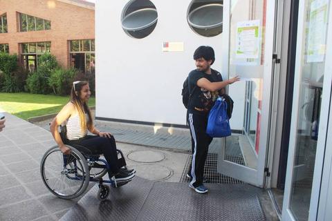 Una niña en silla de ruedas y un joven con mochila están entrando a un edificio.