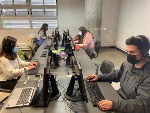 Un grupo de cuatro personas trabaja en computadoras en un ambiente de oficina, usando mascarillas y cuidando el distanciamiento.
