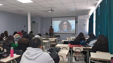 Una clase con alumnos asistiendo a una videoconferencia proyectada en una pantalla.