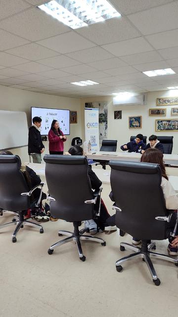Una reunión en una sala con varias personas sentadas, mientras una mujer presenta frente a una pantalla.