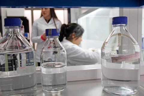 La imagen muestra frascos de laboratorio con líquido en primer plano y dos científicas trabajando en el fondo.