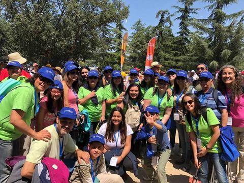 Un grupo grande de personas sonrientes con camisetas verdes y gorras azules disfrutando de un día al aire libre.