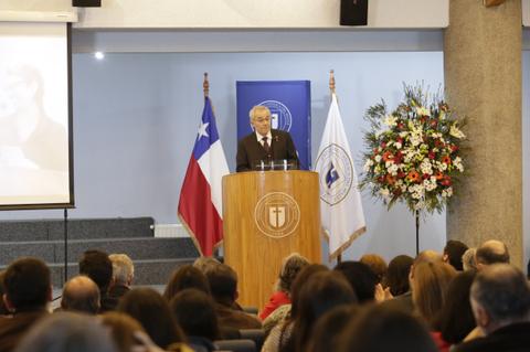 Un hombre habla desde un podio en un evento formal, con una bandera chilena y un público atento en la audiencia.