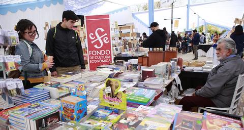 Una feria del libro donde dos personas observan una mesa llena de libros mientras un hombre mayor las atiende.