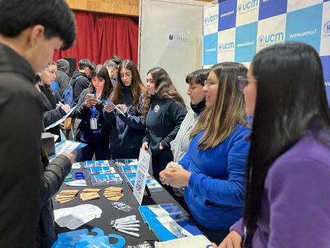 Un grupo de estudiantes interactúa en un stand universitario con diversos materiales promocionales.