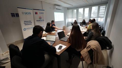 Una reunión de trabajo en una sala con varias personas alrededor de una mesa con computadoras portátiles.