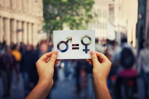 Una persona sostiene un cartel que muestra los símbolos de género masculino y femenino junto a un signo de igualdad en una calle concurrida.