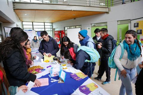 Un grupo de jóvenes interactúa en un evento escolar mientras examinan materiales en una mesa.
