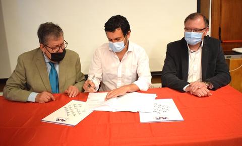 Tres personas se encuentran firmando documentos en una mesa con un mantel rojo.