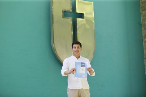 Una persona sostiene un documento frente a una pared con un escudo y una cruz en un fondo azul.