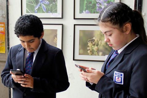 Dos estudiantes en uniforme escolar están usando sus teléfonos móviles mientras miran hacia abajo.