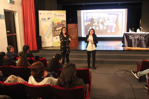 Una presentación en un auditorio con dos mujeres hablando frente a una audiencia sentada.