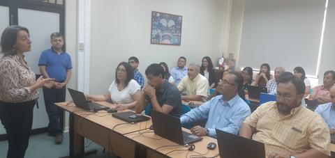 Una presentadora está hablando frente a un grupo de personas en un aula con computadoras portátiles.