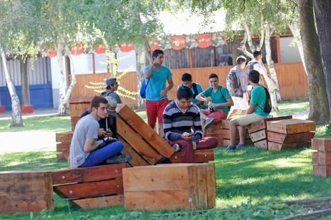 Un grupo de jóvenes se encuentra sentado en un parque en muebles de madera, algunos conversando y otros comiendo.
