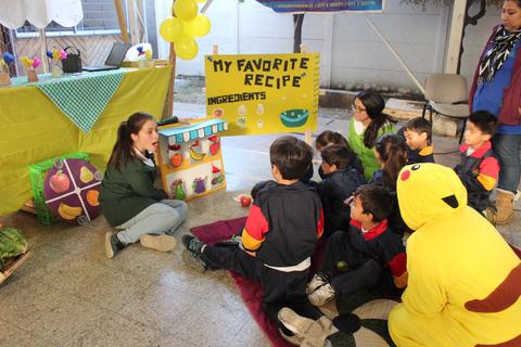 Un grupo de niños escucha atentamente a una persona que presenta una receta en un evento educativo.