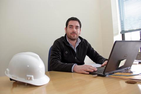 Un hombre sentado frente a una computadora en una oficina, con un casco de seguridad en la mesa.