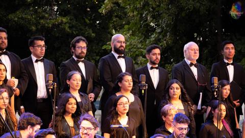 Un grupo de cantantes de pie en un escenario, vestidos formalmente, durante una presentación musical al aire libre.