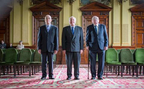 Tres hombres de pie en un salón con sillas verdes vacías.