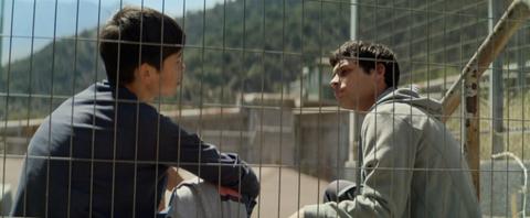 Dos jóvenes conversan a través de una reja, en un entorno montañoso.