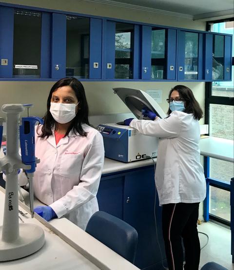 Dos mujeres en un laboratorio, usando mascarillas y batas blancas mientras trabajan con un equipo de análisis.
