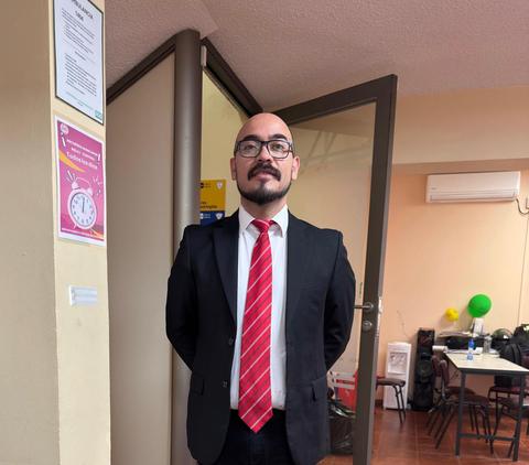 Un hombre de pie con traje y corbata frente a una puerta en un ambiente de oficina.