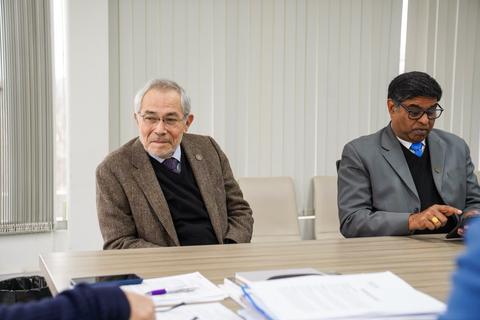 Dos hombres sentados en una reunión, uno con un abrigo de tweed y el otro en traje, en un ambiente moderno.