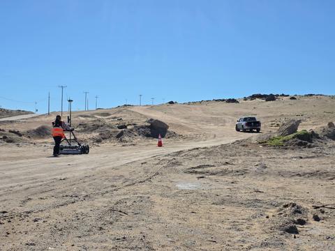 Un trabajador está nivelando un camino en un sector árido mientras un automóvil pasa por el costado.