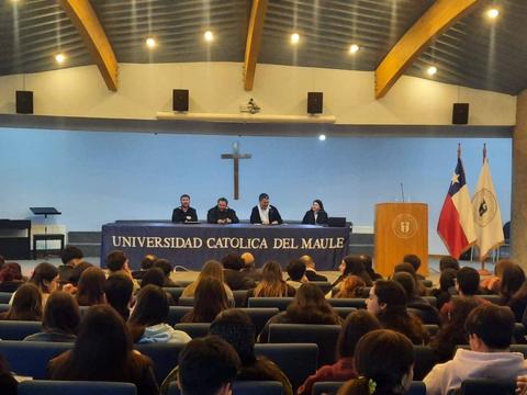 Un grupo de personas sentadas en un auditorio de la Universidad Católica del Maule, con una cruz iluminada al fondo.