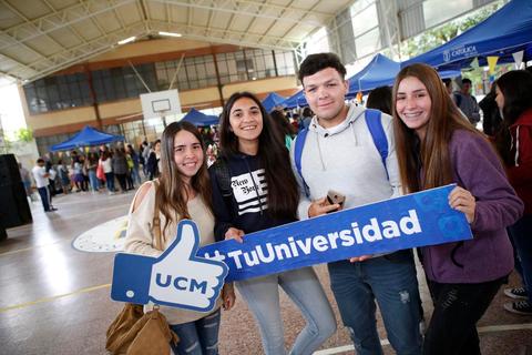 Grupo de cuatro jóvenes sonrientes sosteniendo un cartel de promoción universitaria en un ambiente escolar.