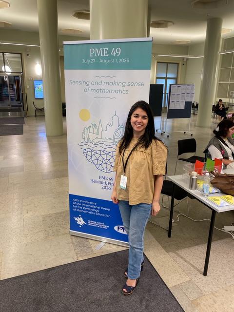 Mujer sonriente posando junto a un banner de una conferencia sobre matemáticas en Finlandia.