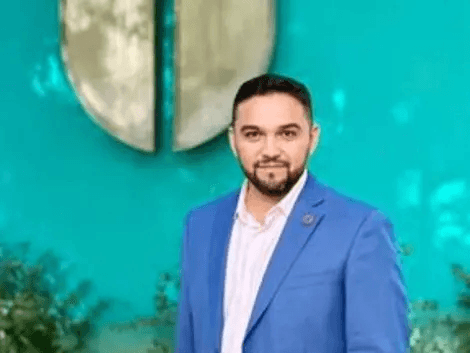 Un hombre con chaqueta azul posando frente a un fondo verde.