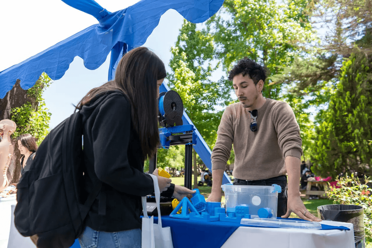 En una feria al aire libre, un joven muestra a una mujer varios objetos de plástico de color azul sobre una mesa.