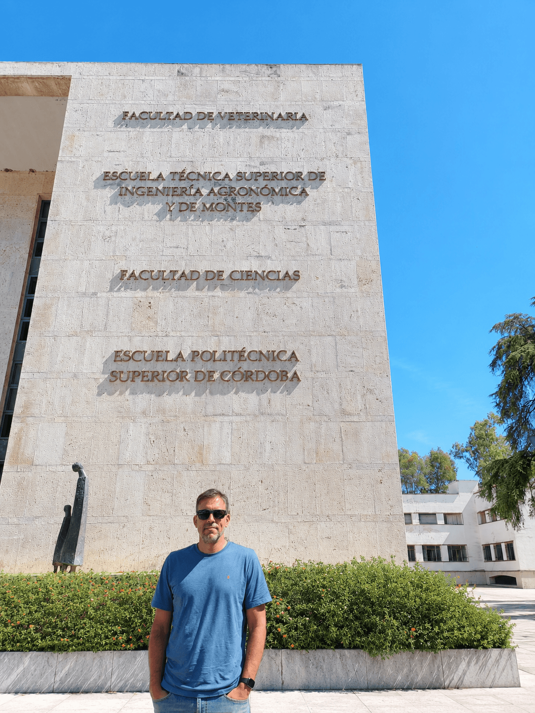 Un hombre posando frente a la fachada de una facultad universitaria en un día soleado.