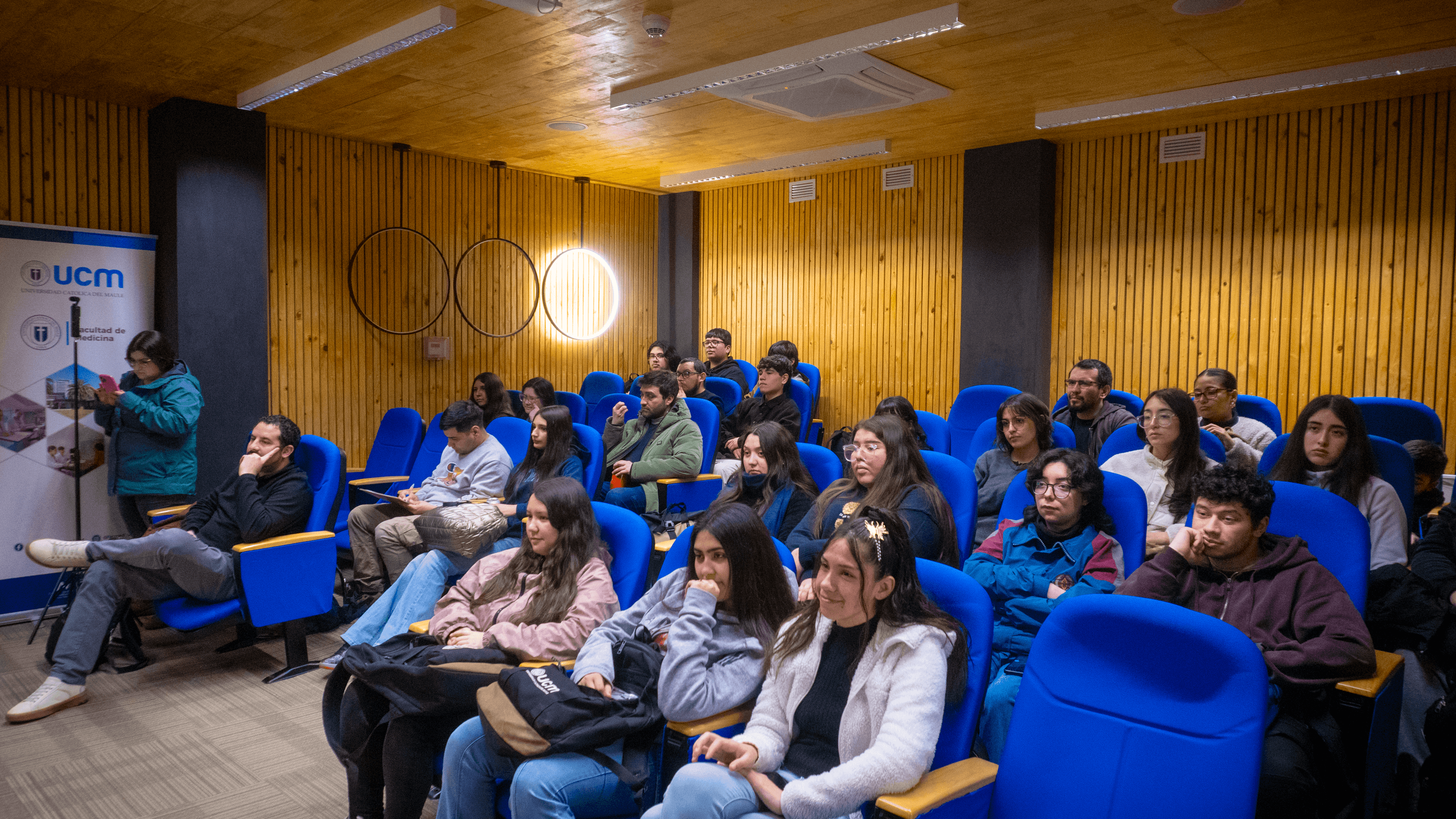 Un grupo de personas sentadas en una sala con asientos azules, escuchando atentamente a un expositor.