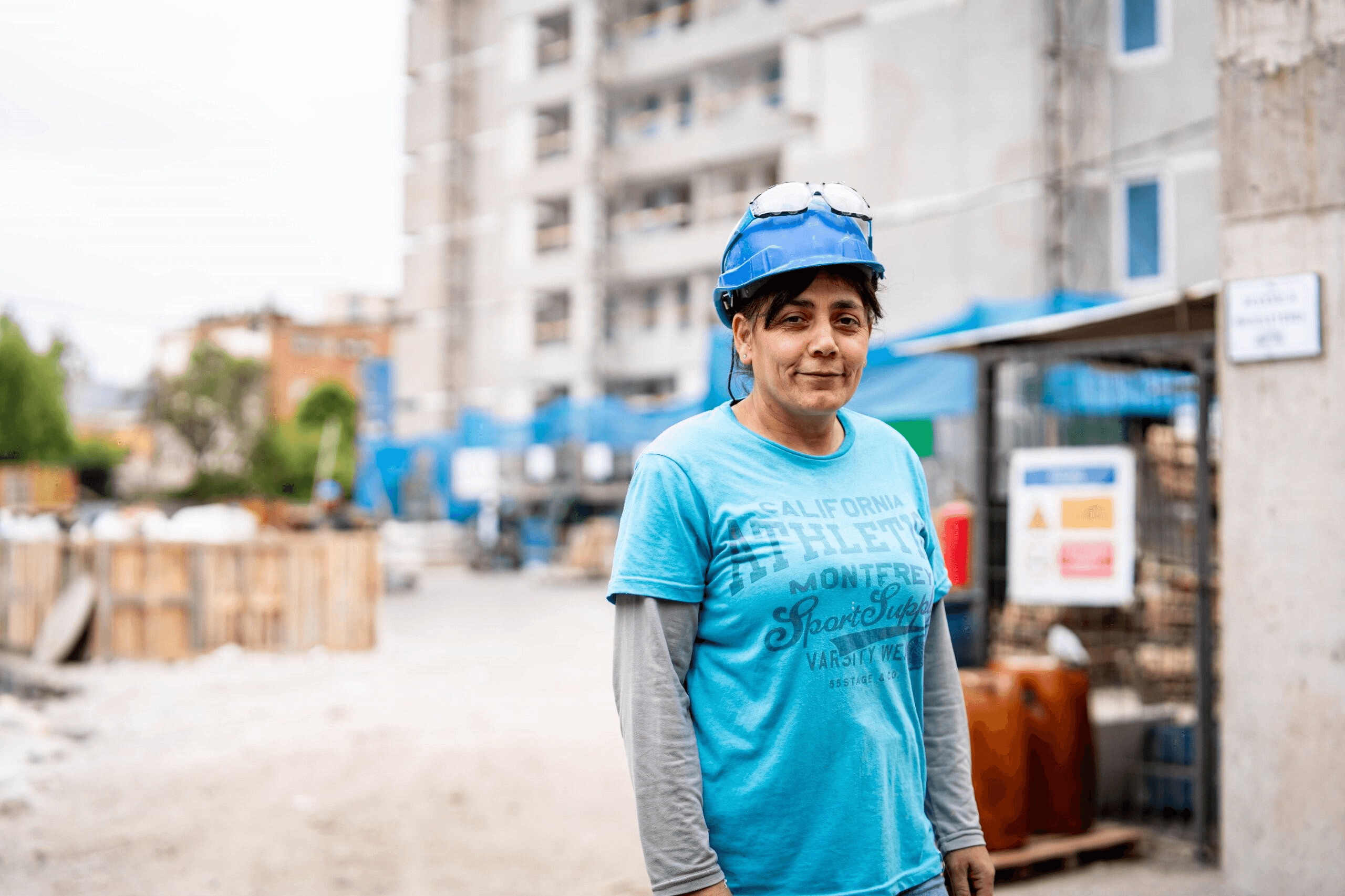Mujer trabajando en una obra de construcción, con casco y un ambiente urbano al fondo.
