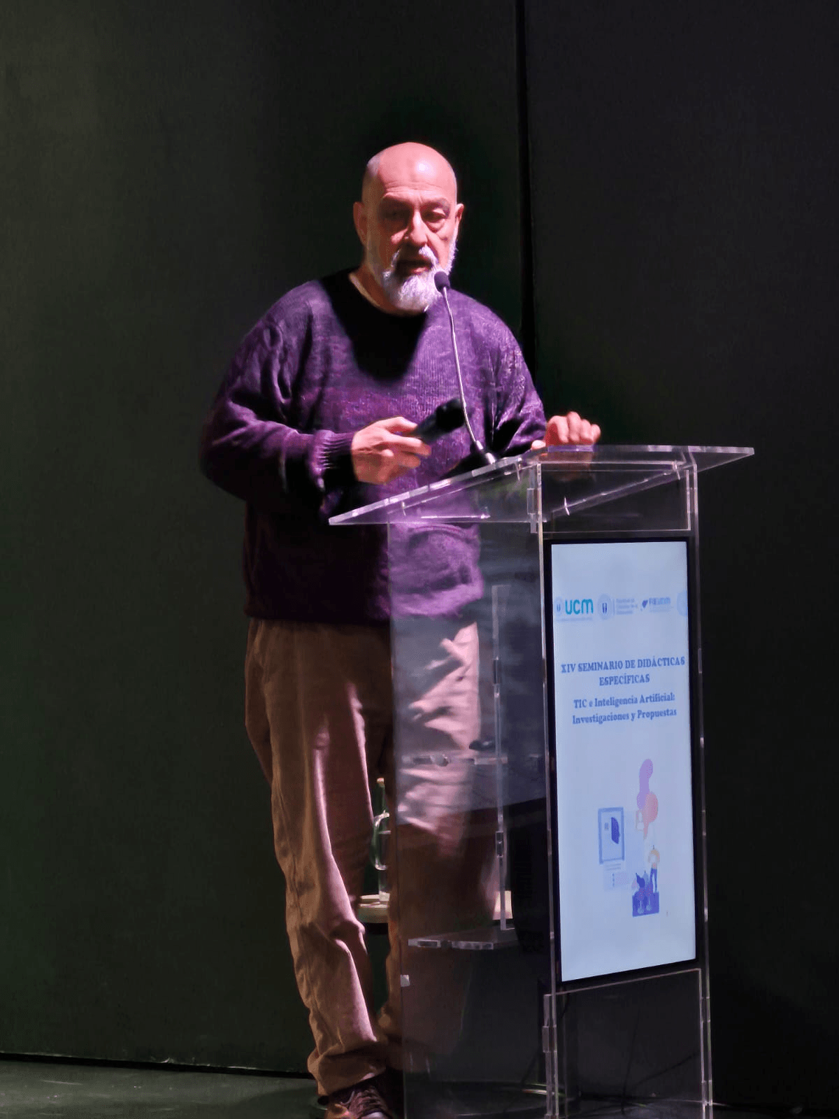 Un hombre con barba presenta en una conferencia, sosteniendo un micrófono frente a un podio.