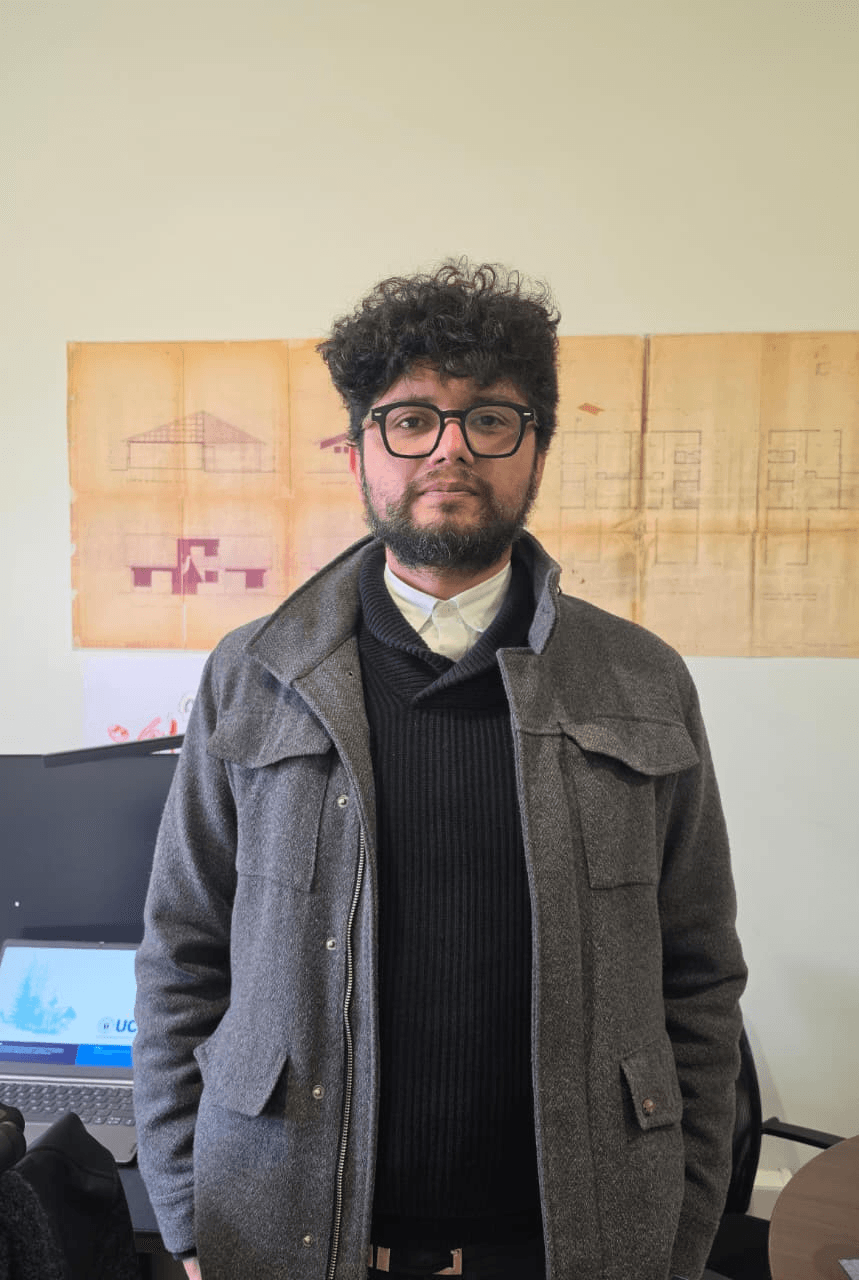 Un caballero posando en una oficina con un fondo de planos arquitectónicos.